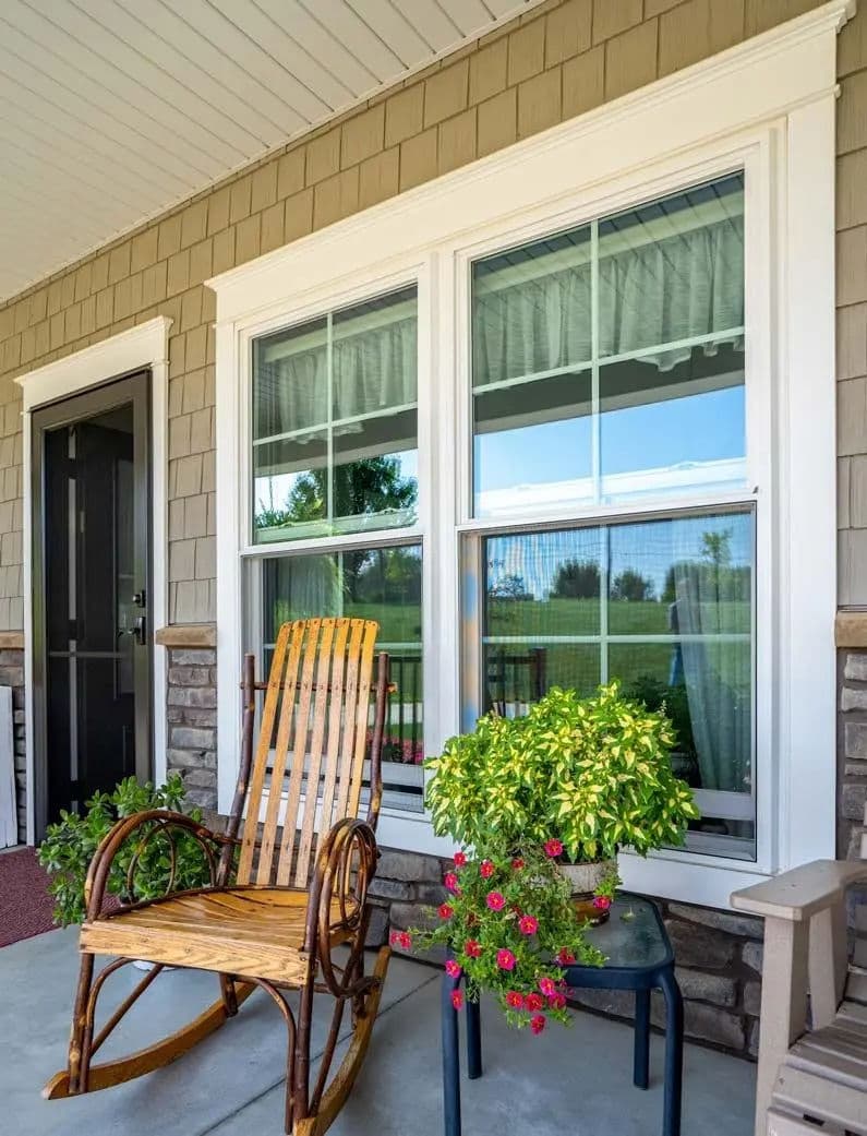 Rocking chair on a porch with potted plants and large windows, inviting outdoor space.